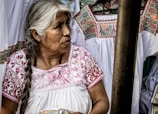 An older woman with long, braided gray hair is wearing a traditional white garment with intricate red embroidery. She is surrounded by similar embroidered garments, suggesting a cultural or artisanal setting.