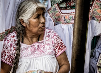 An older woman with long, braided gray hair is wearing a traditional white garment with intricate red embroidery. She is surrounded by similar embroidered garments, suggesting a cultural or artisanal setting.