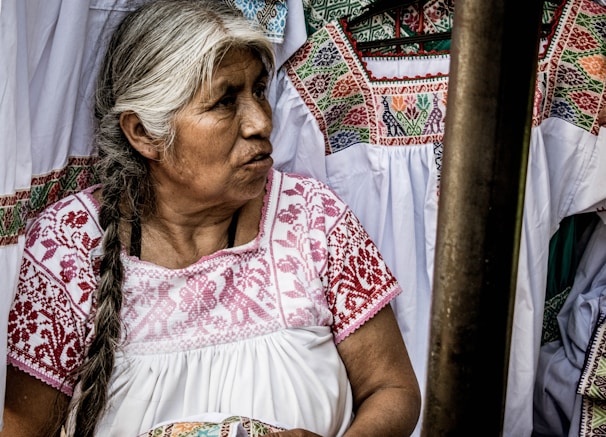An older woman with long, braided gray hair is wearing a traditional white garment with intricate red embroidery. She is surrounded by similar embroidered garments, suggesting a cultural or artisanal setting.