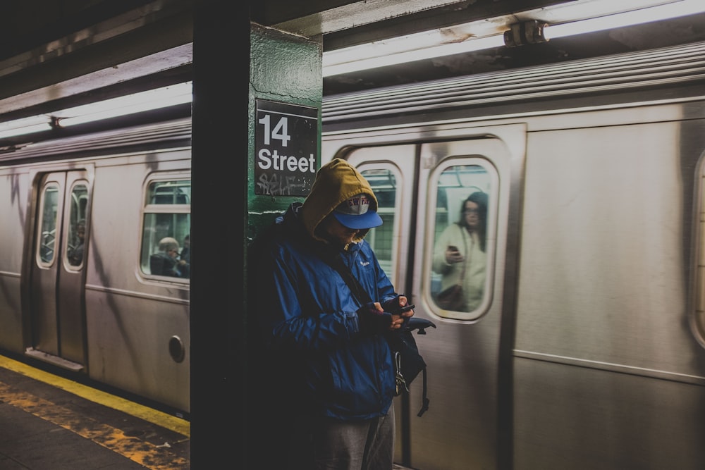 Mann im blauen Kapuzenpullover mit Smartphone im Bahnhof mit Zug in der Nähe