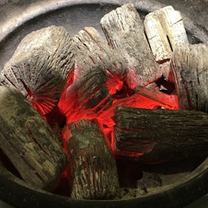 A collection of charred wooden logs with glowing red embers nestled in their crevices. The logs are arranged inside a round metal container, with ash and soot visible on their surfaces.