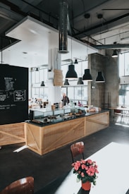 A modern café interior with large windows allowing natural light to flood the space. The counter is made of light wood with a glass display case containing various pastries. Overhead, black pendant lights hang from a high ceiling adorned with exposed ducts. A chalkboard menu is visible on one wall. There are minimalist chairs and tables, one of which has a vibrant pink potted plant. A person stands near the counter, appearing to interact with the barista.