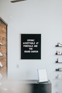 A black sign with white text states, 'CRYING: ACCEPTABLE AT FUNERALS AND THE GRAND CANYON.' The sign is mounted on a white wall. To the right are several small shelves holding cards or books. On the left, part of a wooden wall and additional shelving are visible. Below the sign is a black counter with a sign or book placed on it.