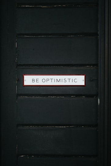 A dark green wall features a rectangular sign with a red border containing the text 'BE OPTIMISTIC' in bold, capital letters.