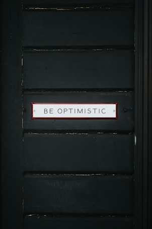 A dark green wall features a rectangular sign with a red border containing the text 'BE OPTIMISTIC' in bold, capital letters.