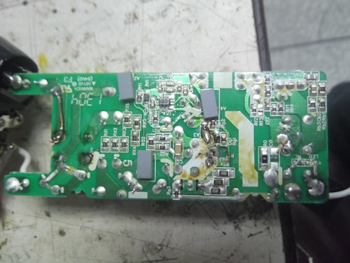 A close-up view of a green printed circuit board (PCB) featuring a variety of silver and black electronic components. There are several connections, solder points, and visible traces along with some text labeling on the surface. The board appears to be resting on a slightly worn, off-white workbench with some signs of use around it.