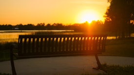 A park bench is silhouetted against a vibrant sunset over a calm body of water. The sun is low on the horizon, casting an orange glow across the landscape. Trees and grass border the scene, creating a peaceful and serene atmosphere.
