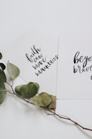 Two white cards with inspirational quotes written in black cursive text are overlapping. One card reads 'faith can move mountains' and the other says 'be your brave'. A dried branch with green leaves is placed beneath the cards, adding a natural touch.