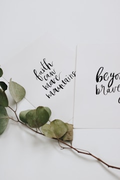 Two white cards with inspirational quotes written in black cursive text are overlapping. One card reads 'faith can move mountains' and the other says 'be your brave'. A dried branch with green leaves is placed beneath the cards, adding a natural touch.