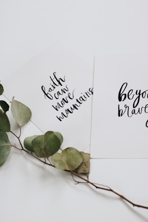 Two white cards with inspirational quotes written in black cursive text are overlapping. One card reads 'faith can move mountains' and the other says 'be your brave'. A dried branch with green leaves is placed beneath the cards, adding a natural touch.