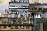 A bakery counter displaying a variety of pastries and desserts in a glass case, including croissants, cookies, and individual cakes. Shelves behind the counter hold jars and bottles, with packaged goods placed beside the display case. The interior features a modern industrial style with tiled walls and dark stone countertops.