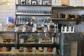 A bakery counter displaying a variety of pastries and desserts in a glass case, including croissants, cookies, and individual cakes. Shelves behind the counter hold jars and bottles, with packaged goods placed beside the display case. The interior features a modern industrial style with tiled walls and dark stone countertops.