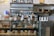 A bakery counter displaying a variety of pastries and desserts in a glass case, including croissants, cookies, and individual cakes. Shelves behind the counter hold jars and bottles, with packaged goods placed beside the display case. The interior features a modern industrial style with tiled walls and dark stone countertops.