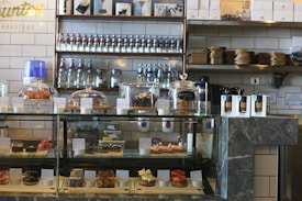 A bakery counter displaying a variety of pastries and desserts in a glass case, including croissants, cookies, and individual cakes. Shelves behind the counter hold jars and bottles, with packaged goods placed beside the display case. The interior features a modern industrial style with tiled walls and dark stone countertops.