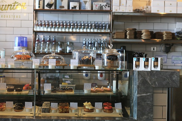 A bakery counter displaying a variety of pastries and desserts in a glass case, including croissants, cookies, and individual cakes. Shelves behind the counter hold jars and bottles, with packaged goods placed beside the display case. The interior features a modern industrial style with tiled walls and dark stone countertops.