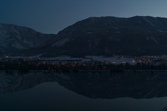 A serene mountain landscape at dusk with a calm lake reflecting the silhouette of the mountains. Small clusters of warm, glowing lights from village homes are scattered across the base of the mountains, creating a peaceful atmosphere.