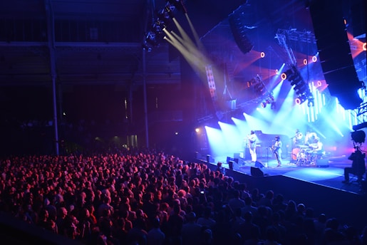 A lively concert scene features a band performing on a brightly lit stage as a large, enthusiastic crowd gathers in the foreground. The stage is bathed in blue and purple lights, with several spotlights and speakers hanging overhead. The venue's architecture is visible in the background, adding depth to the image.