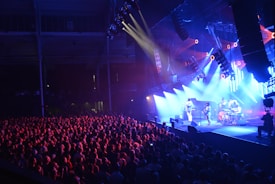 A lively concert scene features a band performing on a brightly lit stage as a large, enthusiastic crowd gathers in the foreground. The stage is bathed in blue and purple lights, with several spotlights and speakers hanging overhead. The venue's architecture is visible in the background, adding depth to the image.