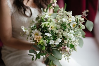 A bouquet of flowers featuring a mix of white, green, and soft pink blooms, elegantly arranged with various foliage. The focus is on the floral arrangement as it is gently held by a person in light-colored attire.