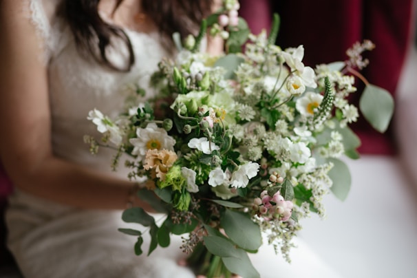 A bouquet of flowers featuring a mix of white, green, and soft pink blooms, elegantly arranged with various foliage. The focus is on the floral arrangement as it is gently held by a person in light-colored attire.