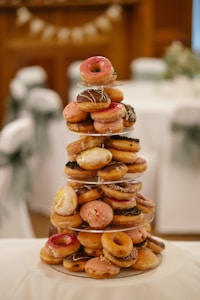 A tower of assorted glazed donuts is neatly stacked on a multi-tiered stand. The donuts vary in toppings, including pink icing, chocolate drizzle, and sprinkles. The setting is elegantly arranged, suggesting a formal or celebratory event with decor in the background, including white tablecloths and a decorative garland.