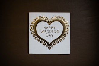 A wedding card featuring a golden heart surrounded by small flower ornaments. The text 'Happy Wedding Day' is elegantly printed in the center. The card is set against a dark background, highlighting its design.