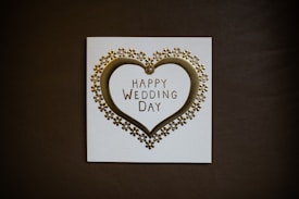 A wedding card featuring a golden heart surrounded by small flower ornaments. The text 'Happy Wedding Day' is elegantly printed in the center. The card is set against a dark background, highlighting its design.
