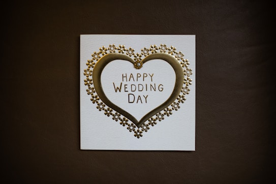 A wedding card featuring a golden heart surrounded by small flower ornaments. The text 'Happy Wedding Day' is elegantly printed in the center. The card is set against a dark background, highlighting its design.