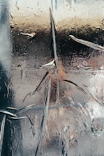 An abstract image featuring a textured surface that appears to be cracked or shattered. The surface has a reflective quality with a mix of metallic and glass-like properties. There are intricate patterns and lines created by the cracks, with some areas reflecting light in various ways.