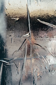 An abstract image featuring a textured surface that appears to be cracked or shattered. The surface has a reflective quality with a mix of metallic and glass-like properties. There are intricate patterns and lines created by the cracks, with some areas reflecting light in various ways.