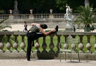 A person in athletic attire is stretching on a stone balustrade in a park setting. A green chair is nearby, and a large stone statue and palm trees can be seen in the background. The scene is peaceful with sparse foot traffic on a sunny day.