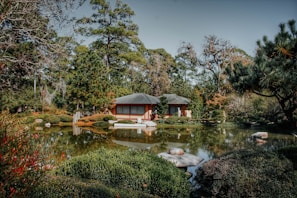 A tranquil Japanese garden featuring a traditional house surrounded by lush greenery and a reflective pond. Large trees and carefully shaped bushes fill the landscape, providing a serene and natural ambiance.