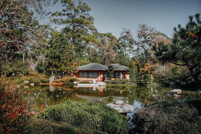 A tranquil Japanese garden featuring a traditional house surrounded by lush greenery and a reflective pond. Large trees and carefully shaped bushes fill the landscape, providing a serene and natural ambiance.