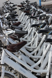 A group of white bicycles arranged in a row, each equipped with black handle grips and baskets. Most seats are brown, and the bikes have a modern design with a clean, organized appearance.