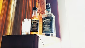 Two bottles of whiskey with distinct labels are placed on a wooden surface. A small, almost full bottle of Jack Daniel's sits beside a cylindrical glass. The background features wooden paneling lit by warm, natural light.