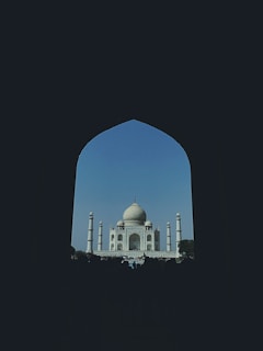 A majestic view of the Taj Mahal framed through a large, dark archway. The iconic white marble mausoleum stands prominently against a clear blue sky, with its intricate architecture and minarets.