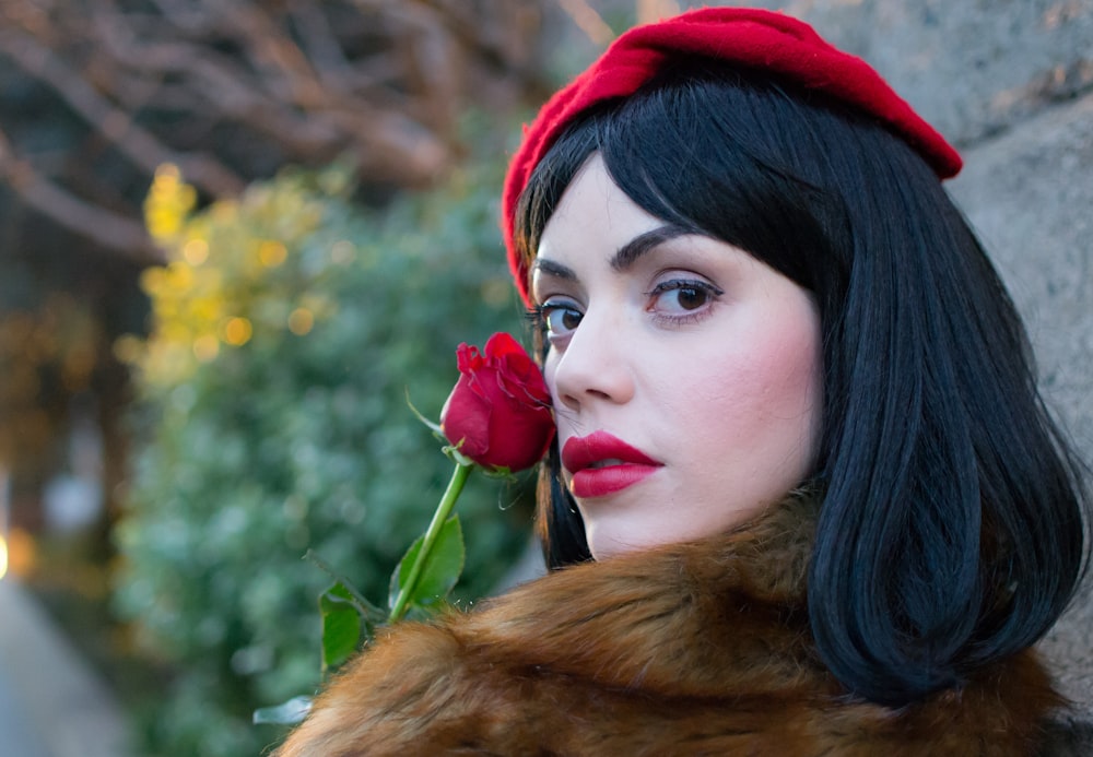 woman wearing brown coat holding red rose near wall during daytime