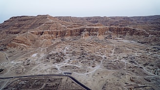 An aerial view of a vast desert landscape with rugged, rocky terrain and scattered ruins. The area features a network of dirt paths and roads. The ground is predominantly sandy with patches of exposed rock. In the foreground, there is a modern road with what appears to be archaeological sites or ruins scattered throughout the area. The background consists of layered, barren hills or mountains with no vegetation visible.