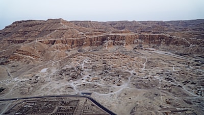 An aerial view of a vast desert landscape with rugged, rocky terrain and scattered ruins. The area features a network of dirt paths and roads. The ground is predominantly sandy with patches of exposed rock. In the foreground, there is a modern road with what appears to be archaeological sites or ruins scattered throughout the area. The background consists of layered, barren hills or mountains with no vegetation visible.