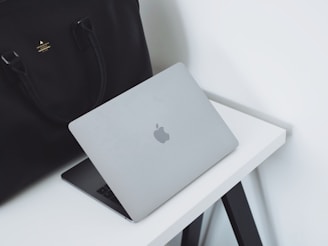 A sleek silver laptop with an Apple logo partially open on a white desk. Beside the laptop is a large black leather bag with handles. The setup is minimalist and professional, placed against a plain white background.