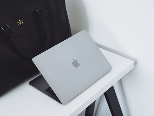 A sleek silver laptop with an Apple logo partially open on a white desk. Beside the laptop is a large black leather bag with handles. The setup is minimalist and professional, placed against a plain white background.