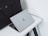 A sleek silver laptop with an Apple logo partially open on a white desk. Beside the laptop is a large black leather bag with handles. The setup is minimalist and professional, placed against a plain white background.