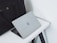 A sleek silver laptop with an Apple logo partially open on a white desk. Beside the laptop is a large black leather bag with handles. The setup is minimalist and professional, placed against a plain white background.