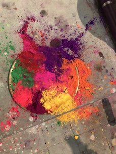 A variety of brightly colored powders including red, yellow, green, pink, and purple are scattered on a concrete surface. The colors create a vibrant and dynamic composition, with a circular plate partially visible beneath the powders.