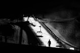 A monochrome night scene captures a snow-covered slope illuminated by lights. A ski lift is visible in the foreground, with silhouettes of people and chairs. The surrounding area is dark, highlighting the glow from the well-lit slope.