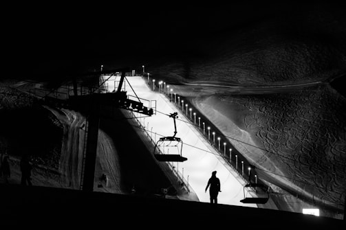 A monochrome night scene captures a snow-covered slope illuminated by lights. A ski lift is visible in the foreground, with silhouettes of people and chairs. The surrounding area is dark, highlighting the glow from the well-lit slope.