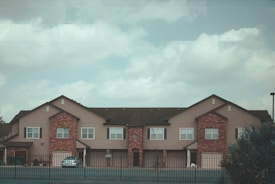 A row of connected residential buildings features brick facades and beige siding, topped with a dark shingle roof. The structures have multiple windows and garage doors, surrounded by a fenced area in the foreground, under a cloudy sky.