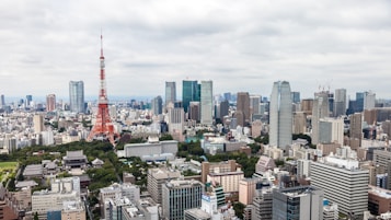 A vast urban cityscape featuring numerous skyscrapers and the prominent red and white structure of Tokyo Tower. The skyline is densely packed with a mix of modern high-rise buildings and older structures. Green spaces are visible, providing contrast to the urban environment. The sky is overcast, adding a muted tone to the scene.