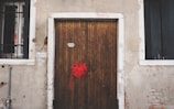 A wooden door with vertical planks is centered between two windows with black metal bars. A vibrant red ribbon is tied to the door handle. The building exterior features an aged, weathered appearance with cracked plaster and exposed bricks. Above the door, a possible address number '9825' is visible, and to the left of the door, there is a street sign labeled 'B AVON'.