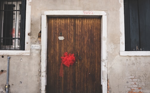 A wooden door with vertical planks is centered between two windows with black metal bars. A vibrant red ribbon is tied to the door handle. The building exterior features an aged, weathered appearance with cracked plaster and exposed bricks. Above the door, a possible address number '9825' is visible, and to the left of the door, there is a street sign labeled 'B AVON'.